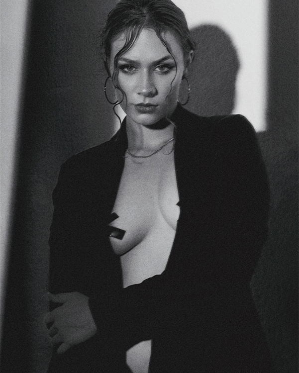 Fotografía editorial modelo sentada en una silla metálica, vestida con chaqueta color café un camisa y pantalón blanco.